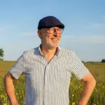 Ulrich Watermann steht lächelnd auf einer bunten Sommerblumenwiese mit Klatschmohn, Lupinen und Hahnenfuß im Sonnenlicht – ein Moment gespürter Stille und Präsenz.